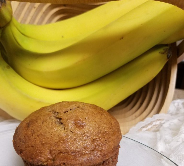 A freshly baked banana bread muffin in front of a bunch of yellow bananas.